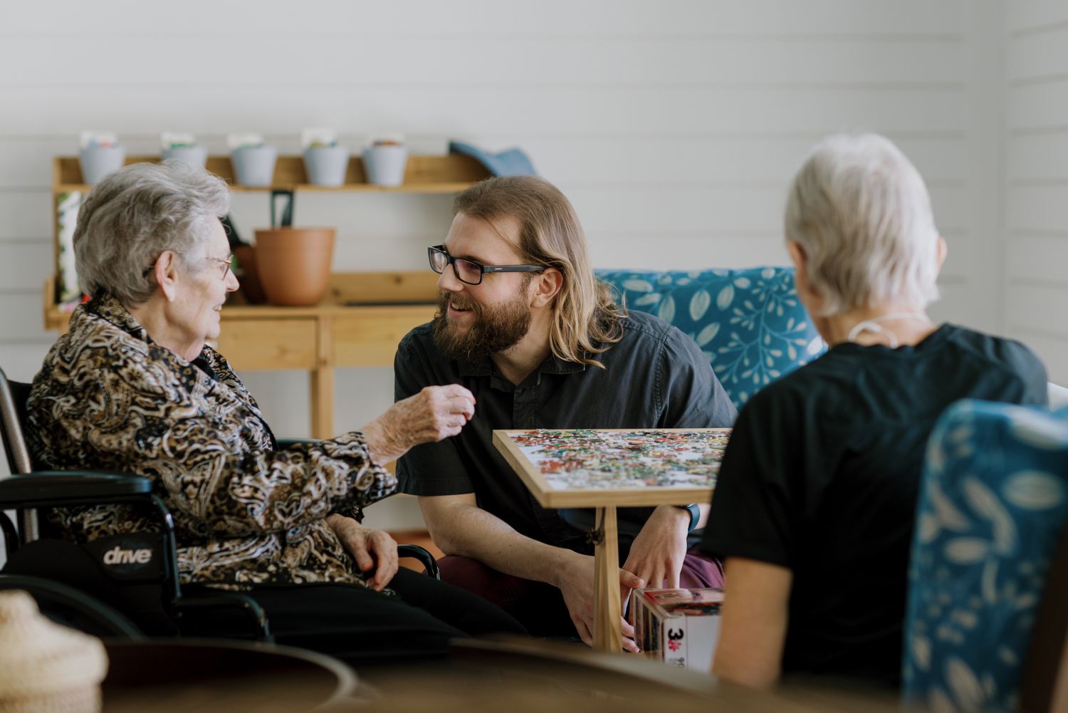 Young man with glasses engaging warmly with two elderly women over a puzzle at a care facility.