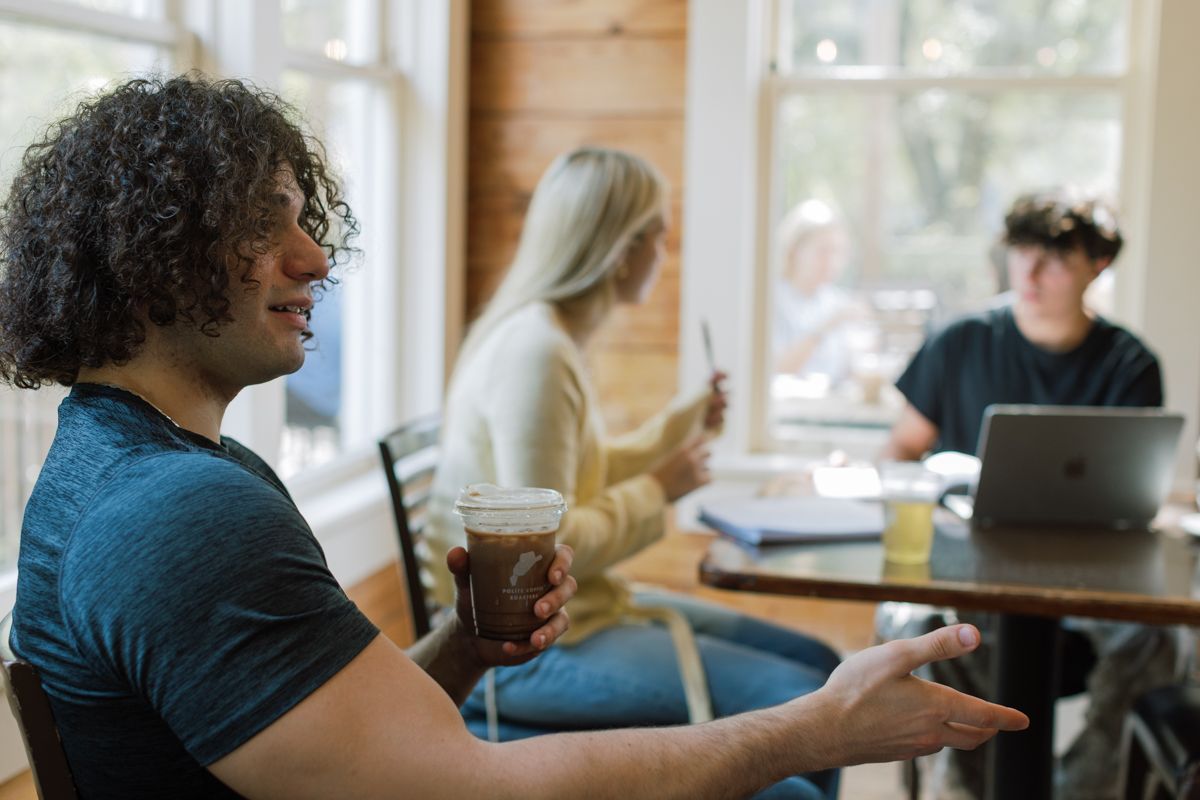 Young man with curly hair holding iced coffee gesturing while talking with friends at a cozy cafe.