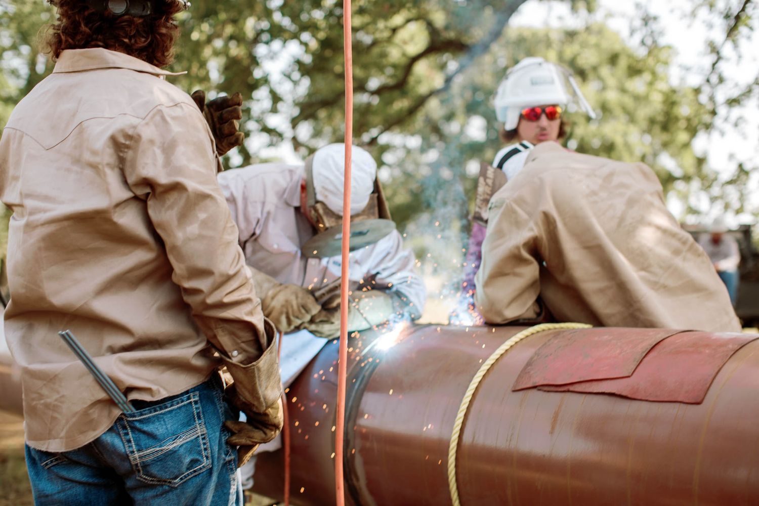 Workers in protective gear welding a large metal pipe outdoors, with sparks flying during an industrial welding operation.