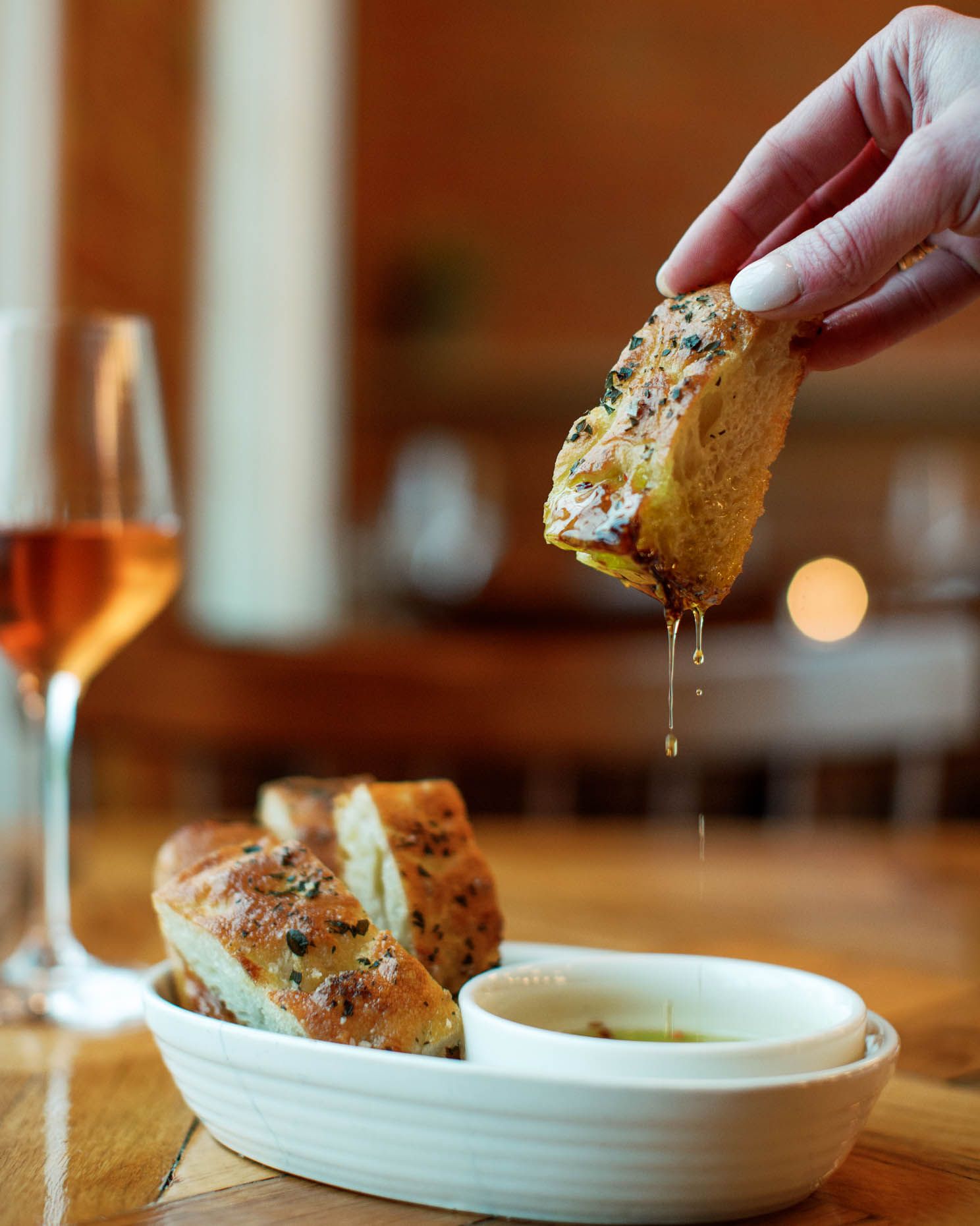 Hand dipping herb-crusted bread into olive oil beside a glass of rosé wine at a rustic restaurant table.