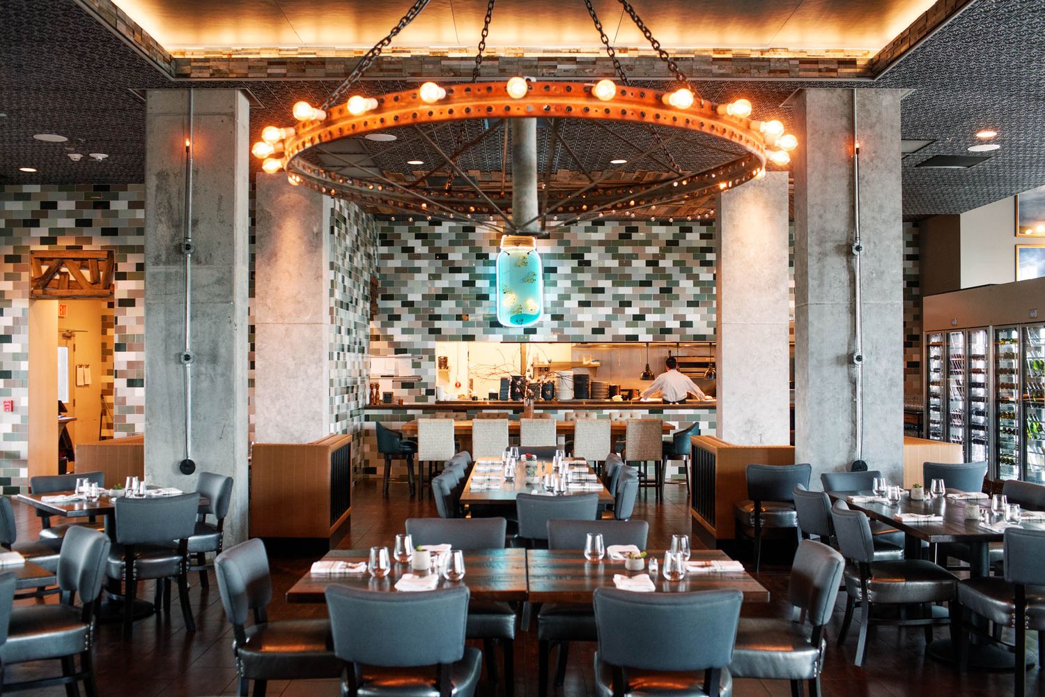Elegant restaurant interior with round chandelier, gray chairs, set tables, and mosaic tile backsplash near open kitchen.