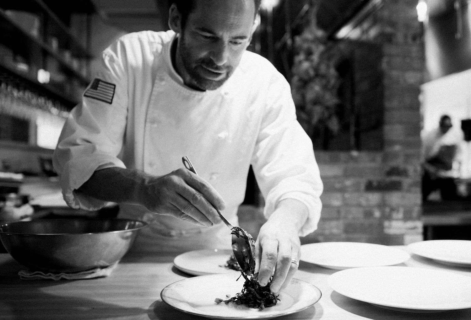 Chef in white coat carefully plating a gourmet dish in a restaurant kitchen, black and white photo.