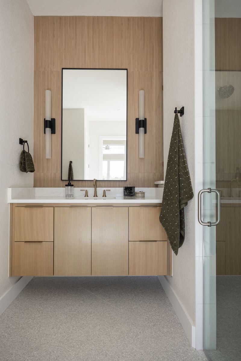 Floating wood vanity with reeded accent wall, black sconces, rectangular mirror, and terrazzo flooring in modern bathroom.