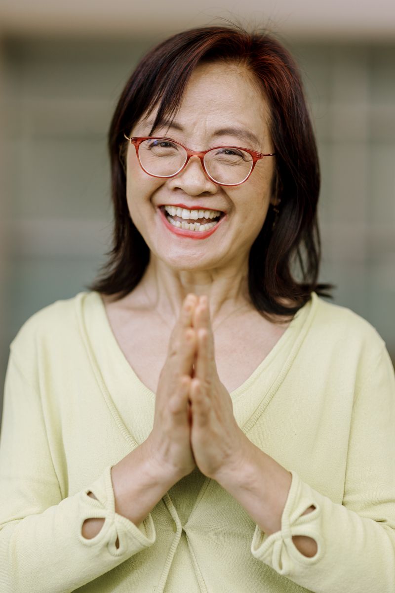 Smiling middle-aged Asian woman with red glasses wearing yellow top, making prayer gesture with hands pressed together.