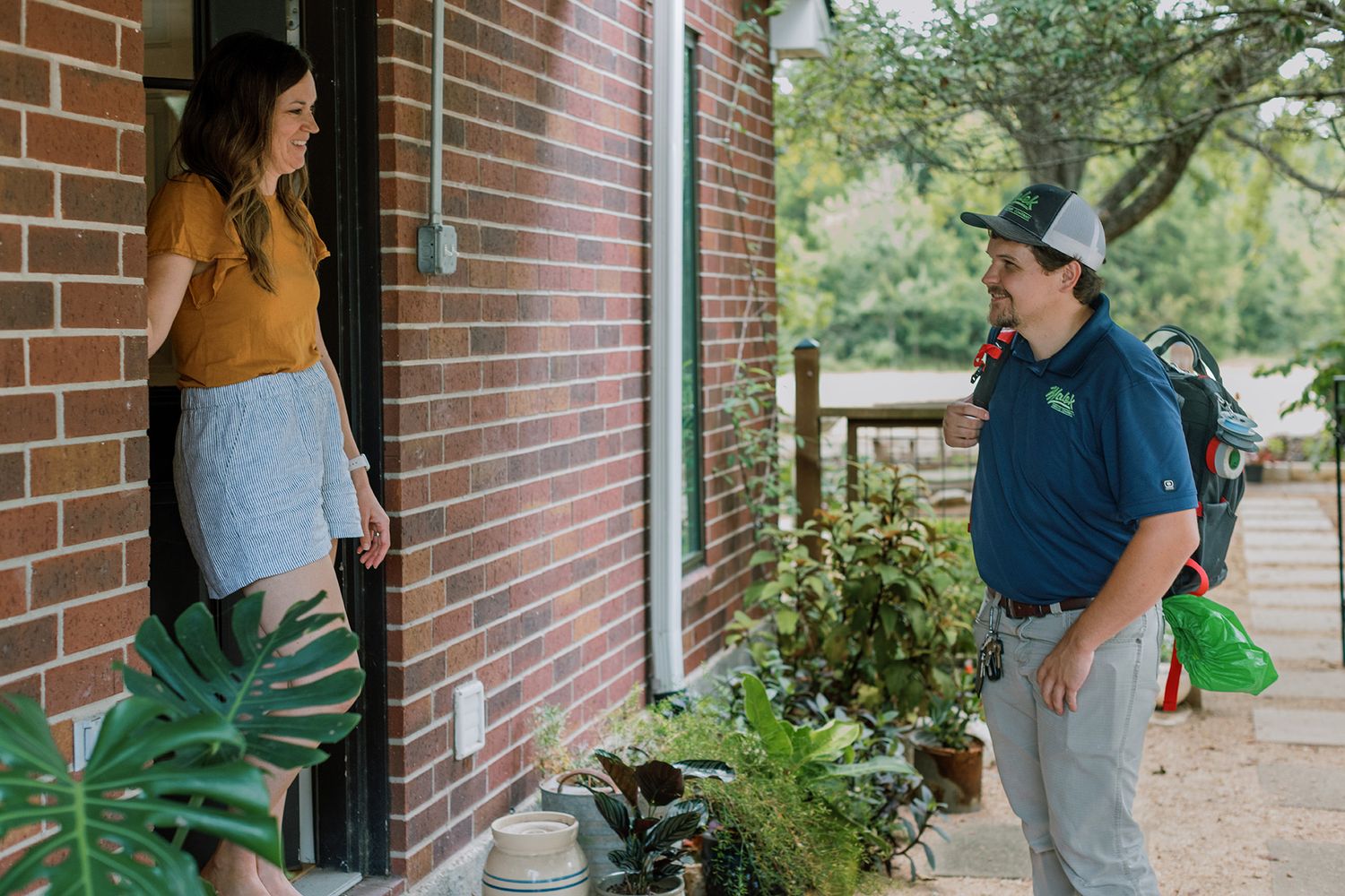 Service technician with backpack greeting a female homeowner at her brick home front door.