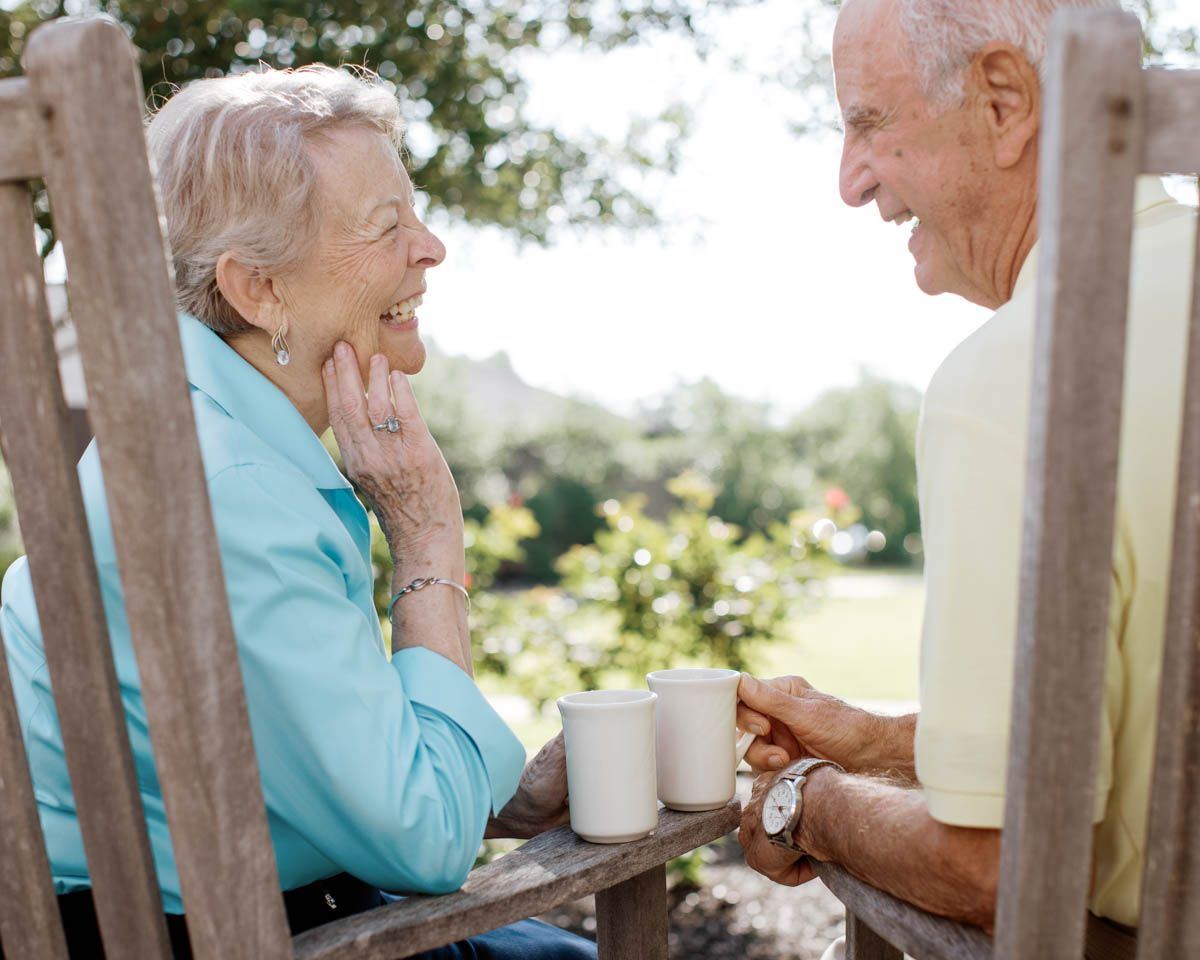 Elderly couple sharing coffee outdoors, smiling and holding hands on a wooden bench surrounded by greenery.