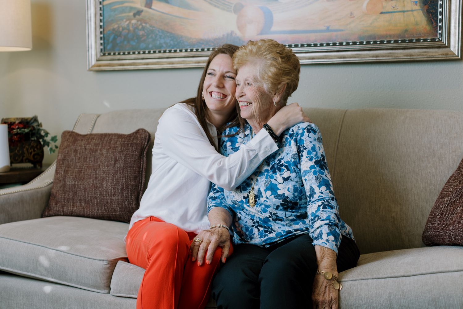 A younger woman hugs an elderly woman on a couch, both smiling warmly in a cozy living room.