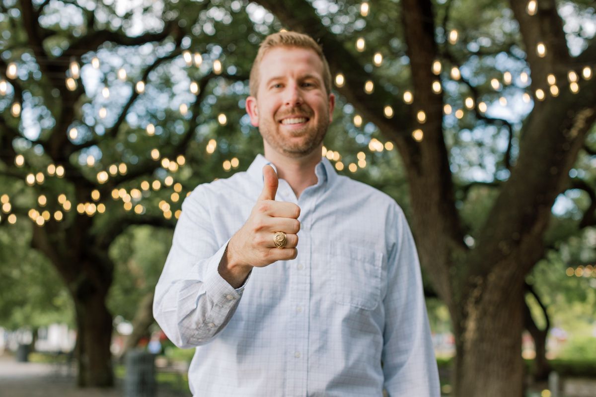 Smiling man giving thumbs up outdoors under trees with string lights in background.