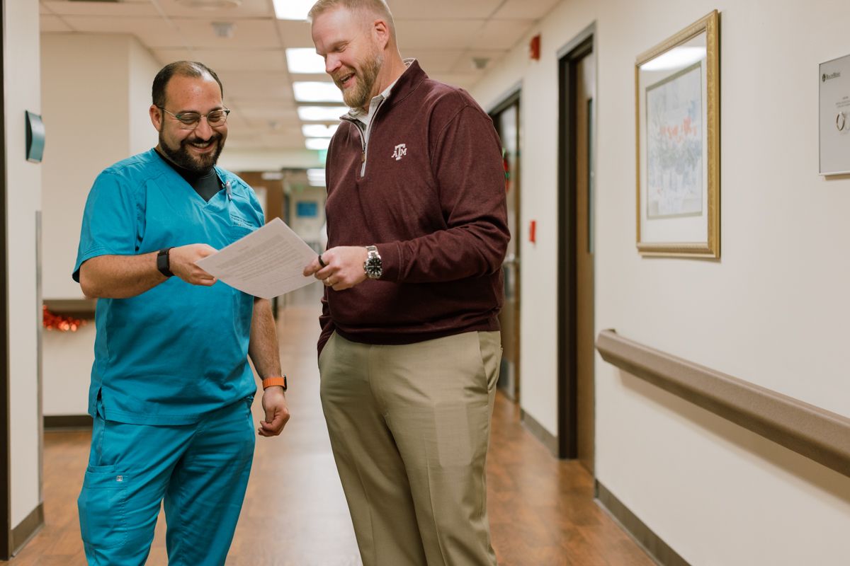 Two male healthcare professionals review a document together while walking in a hospital corridor.