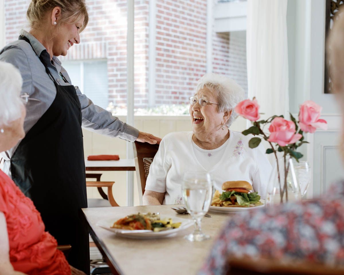 Caregiver serving meals to happy elderly women dining together at a bright, cheerful assisted living facility.