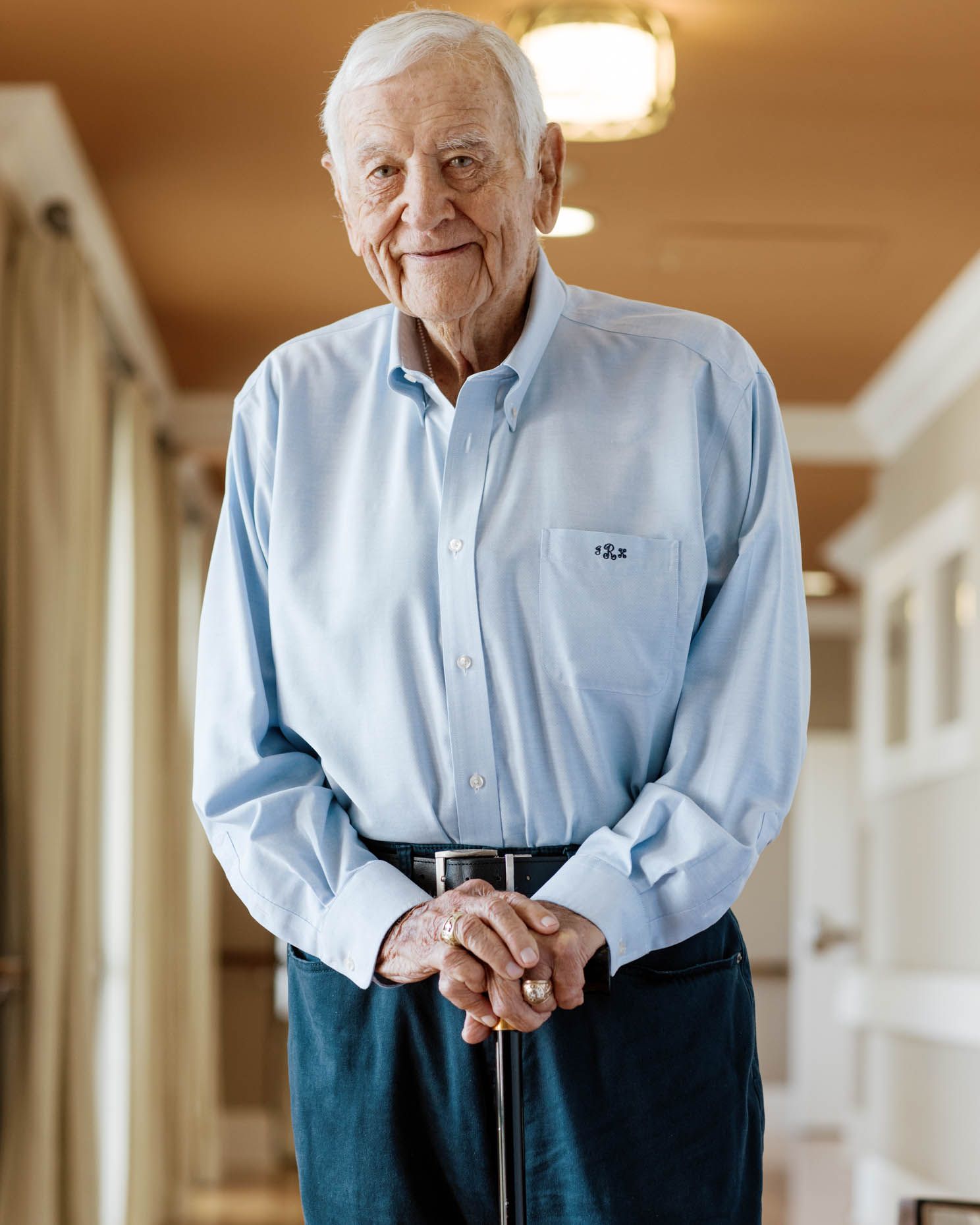Elderly man smiling in a light blue shirt, standing with a cane in a bright hallway.