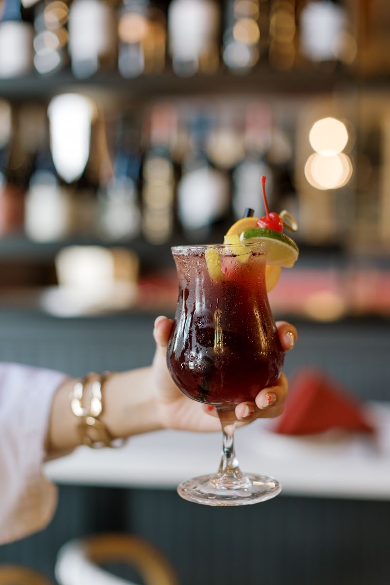 Hand holding a dark red cocktail garnished with cherry and lime slice, with a blurred bar background.