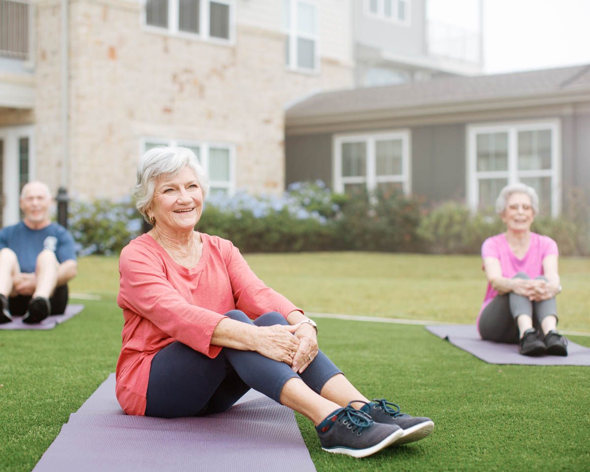 Senior adults doing outdoor yoga on mats, smiling and stretching on green grass near a residential building.