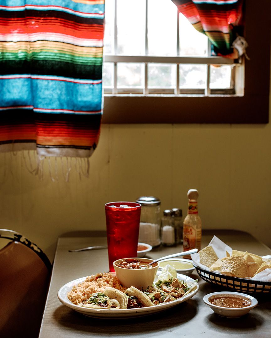 Mexican restaurant table with tacos, chips, salsa, and a red drink near a colorful serape curtain.