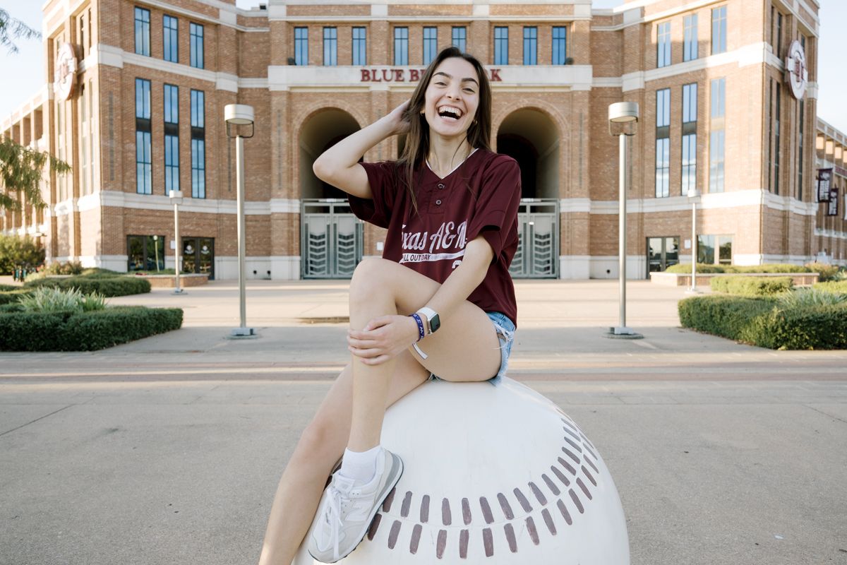 A smiling student sits on a large baseball sculpture outside Kyle Field at Texas A&M.