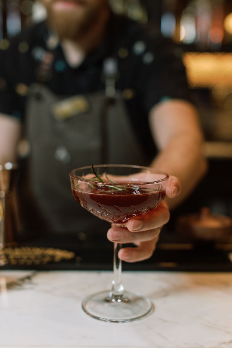 Bartender presenting a dark red cocktail garnished with rosemary in an elegant coupe glass.