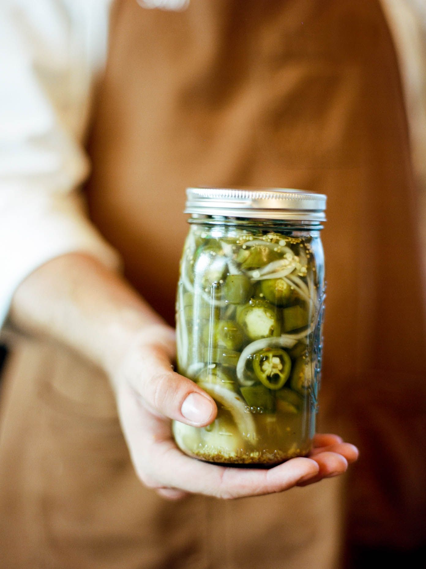 Person in brown apron holding a mason jar filled with pickled jalapenos and onions.