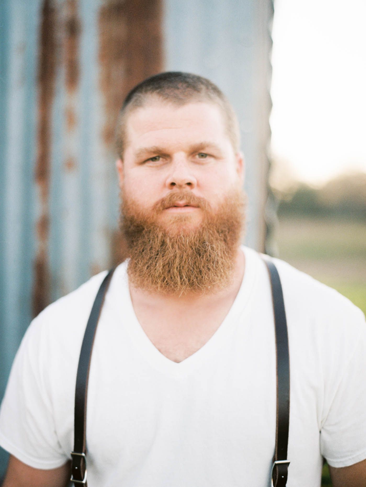 Bearded man with short hair wearing a white v-neck shirt and suspenders standing outdoors near a rustic metal wall.