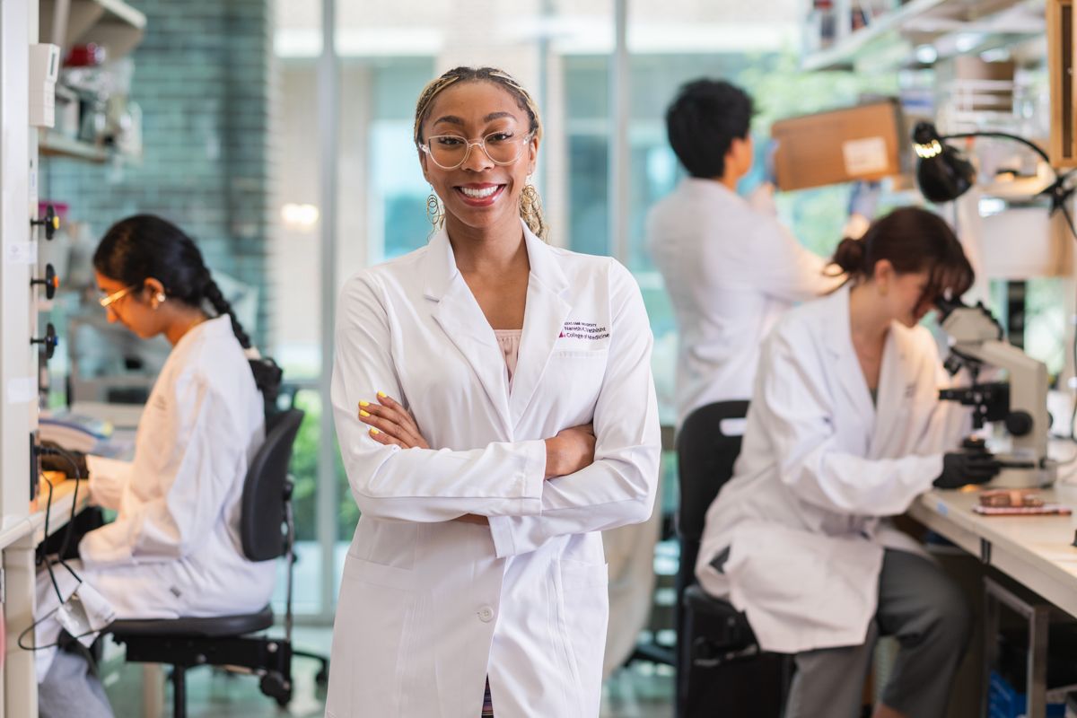 A confident female scientist in a white lab coat stands with arms crossed in a research lab.