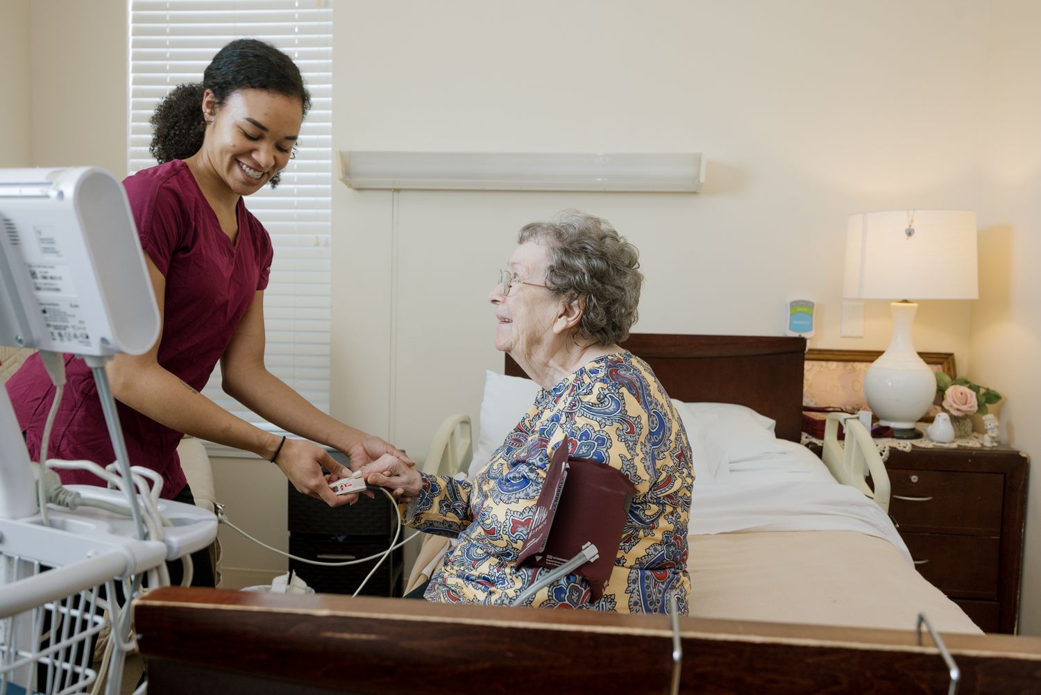 A smiling nurse in red scrubs assists an elderly woman with medical equipment in a cozy care facility room.