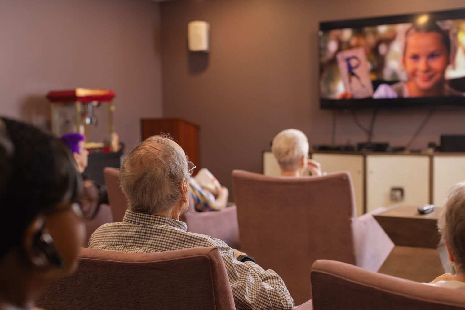 Elderly residents watching a movie on a large TV screen in a cozy senior living community theater room.