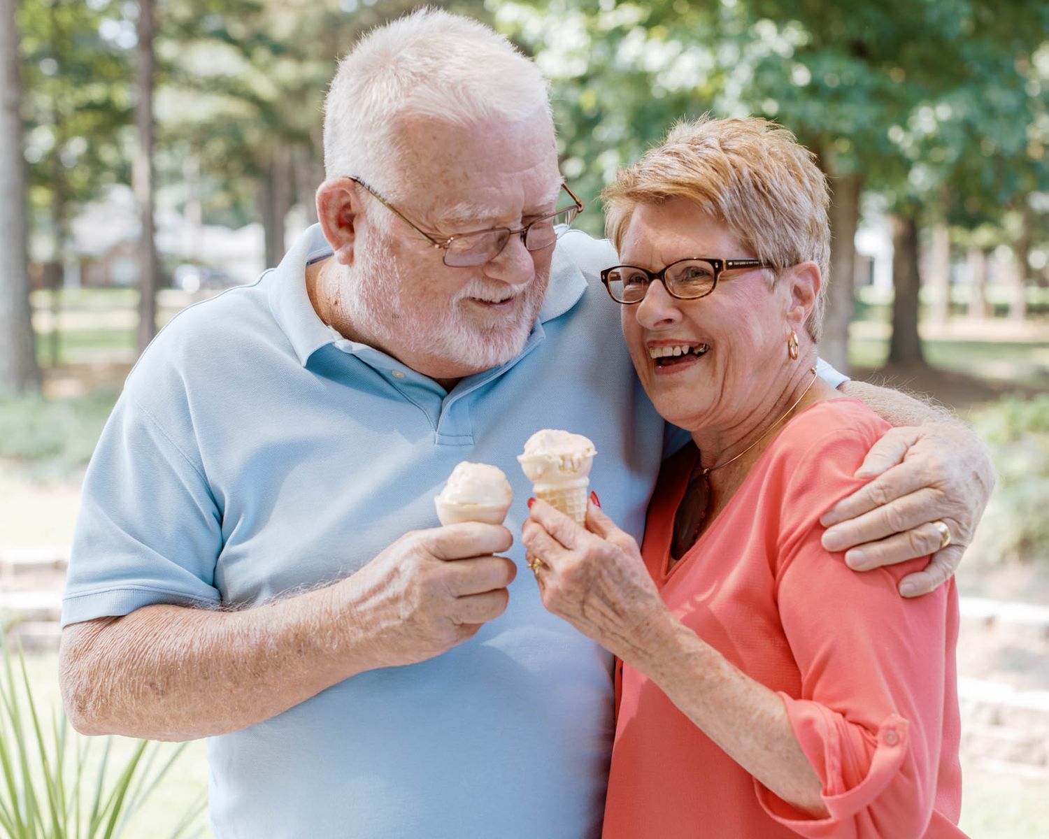 Happy elderly couple sharing ice cream cones outdoors, laughing together in a park setting.