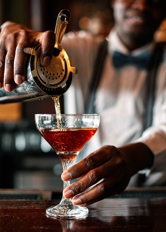 Bartender in bow tie pouring a cocktail into a crystal coupe glass at an upscale bar.