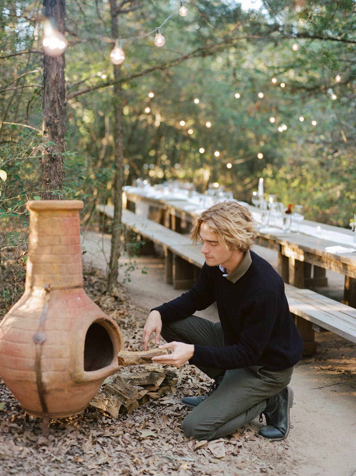 A young man with blonde hair tends to a clay chiminea fireplace outdoors near a long wooden dining table strung with lights.