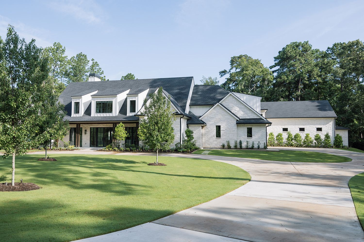Modern farmhouse exterior with white brick, black windows, mixed rooflines, and lush green landscaping.