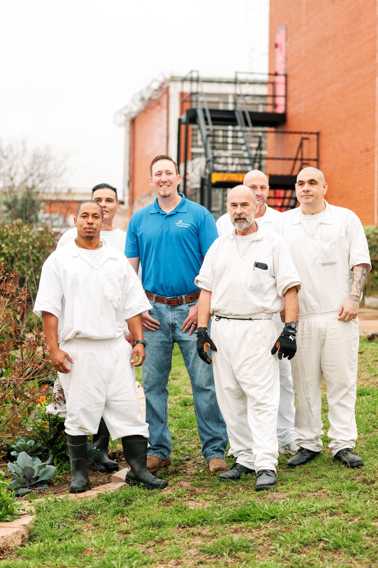 Group of six men in white uniforms and blue polo shirt posing outdoors near a brick building garden.