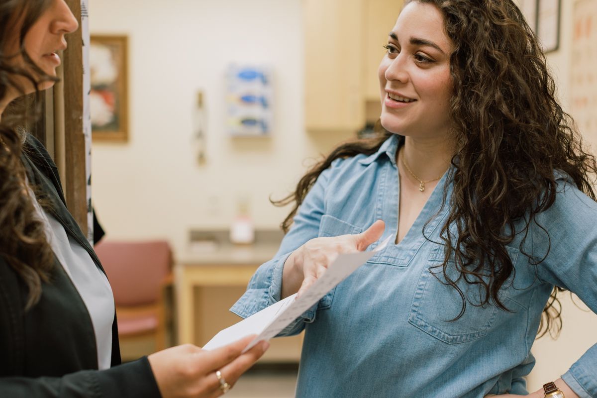 A curly-haired female healthcare worker smiles while handing documents to a colleague in a clinic.