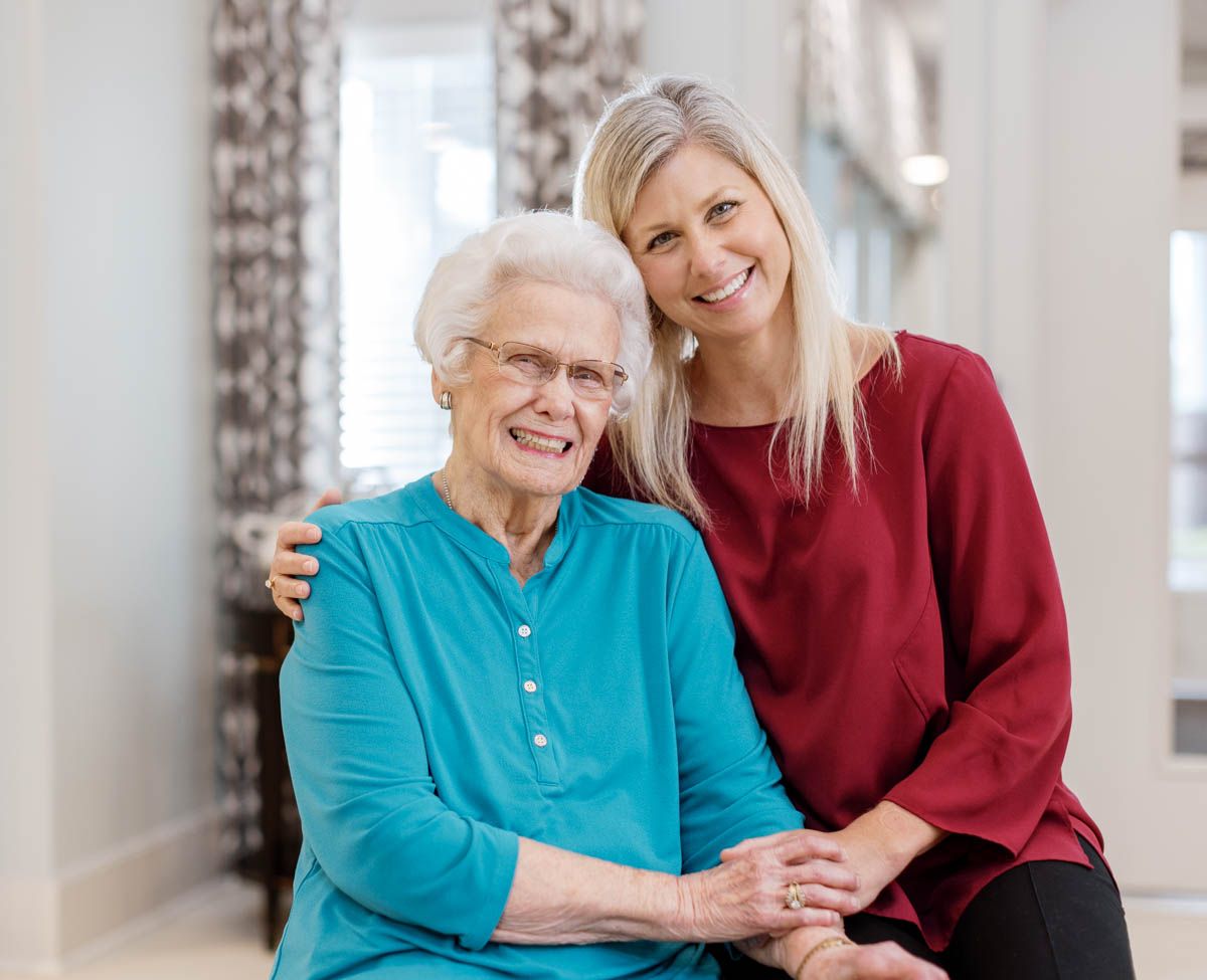 Two women smiling together indoors, an elderly woman seated and a younger blonde woman standing behind her.
