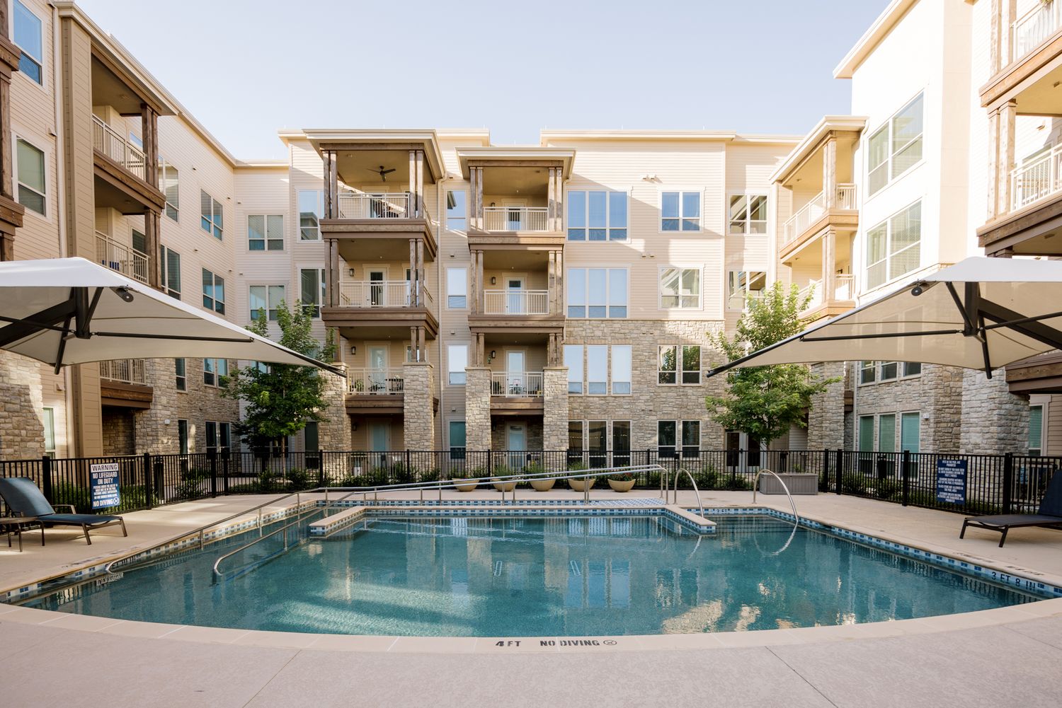 Luxury apartment complex with a sparkling outdoor pool, lounge umbrellas, and multi-story stone-accented buildings.