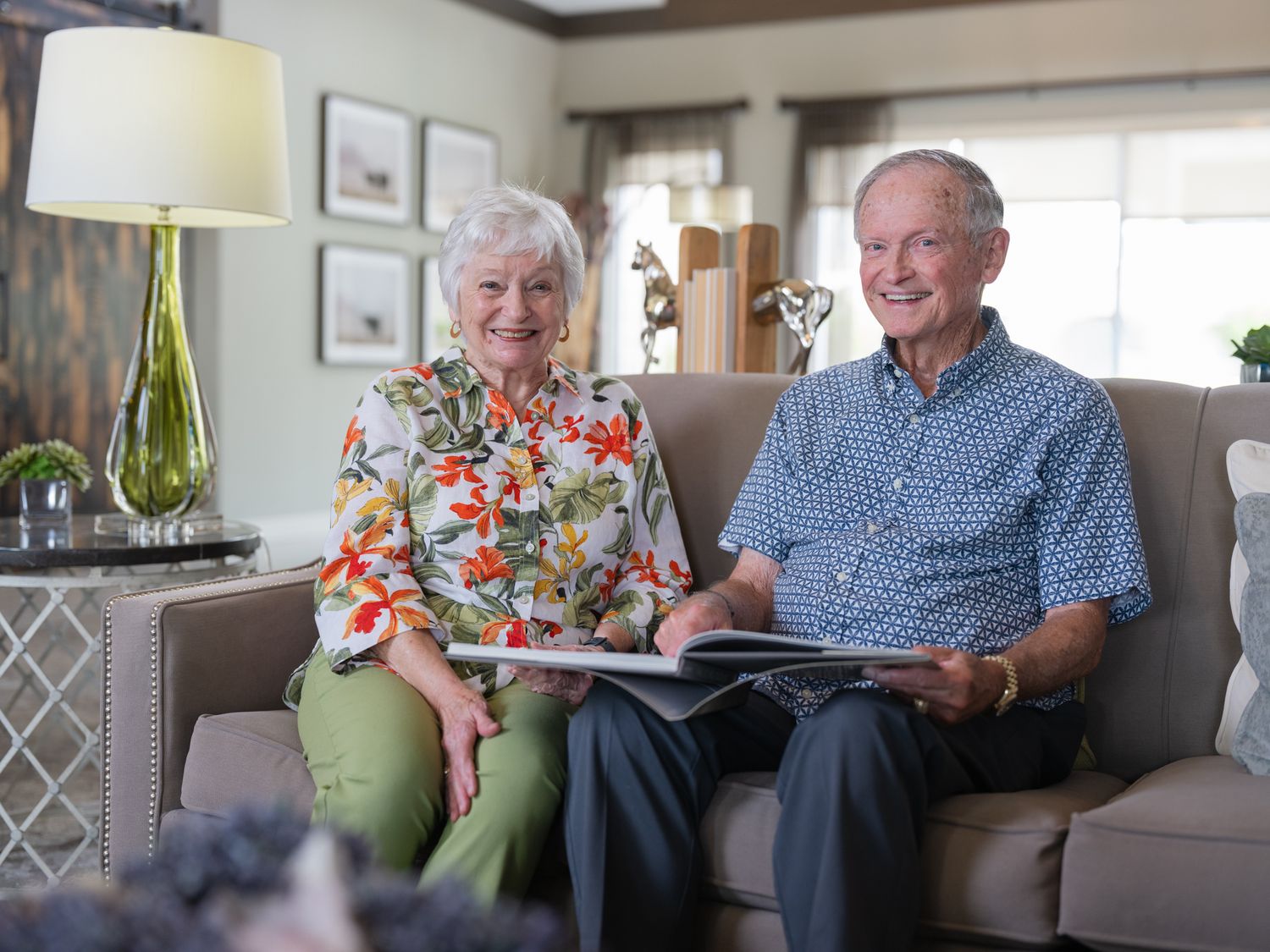 Elderly couple smiling and looking at a photo album together on a couch in a cozy living room.