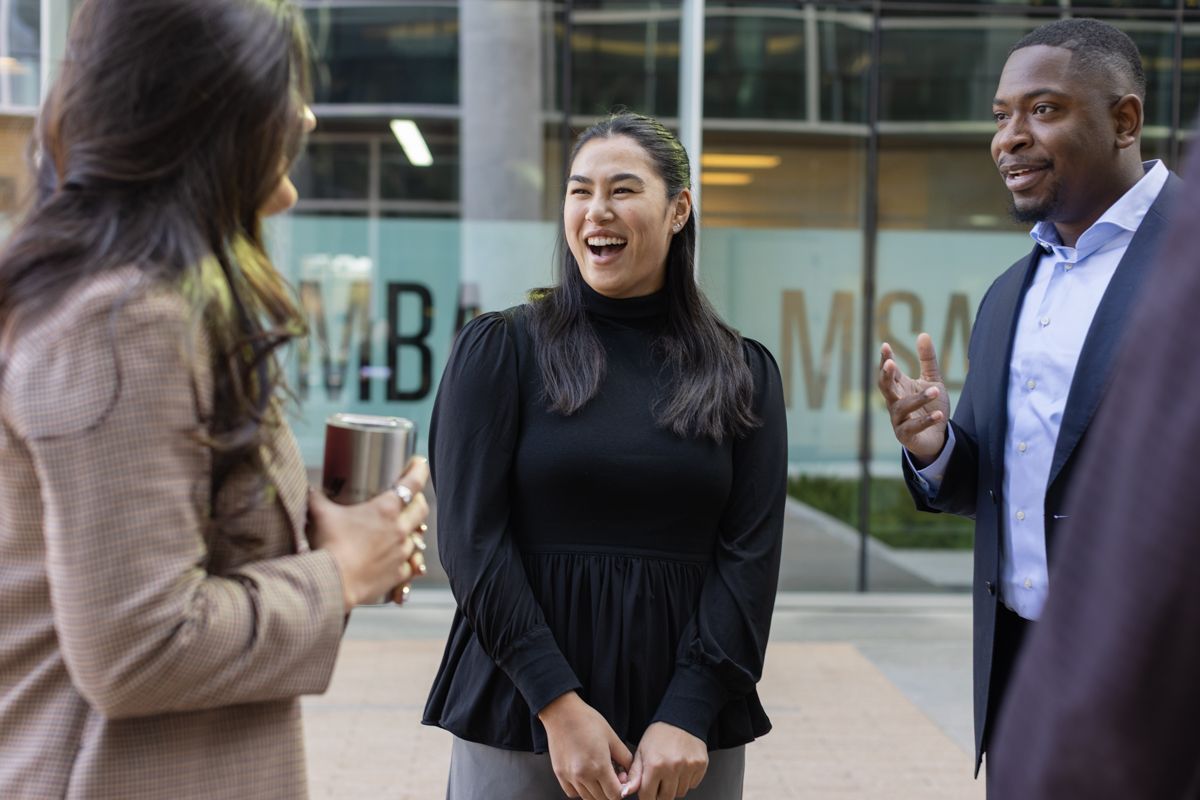 Three diverse colleagues laugh and converse outside a modern MBA program building.