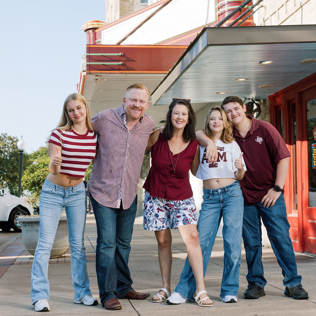 A cheerful group of five people posing together outside a theater on a sunny day, dressed in casual summer attire.