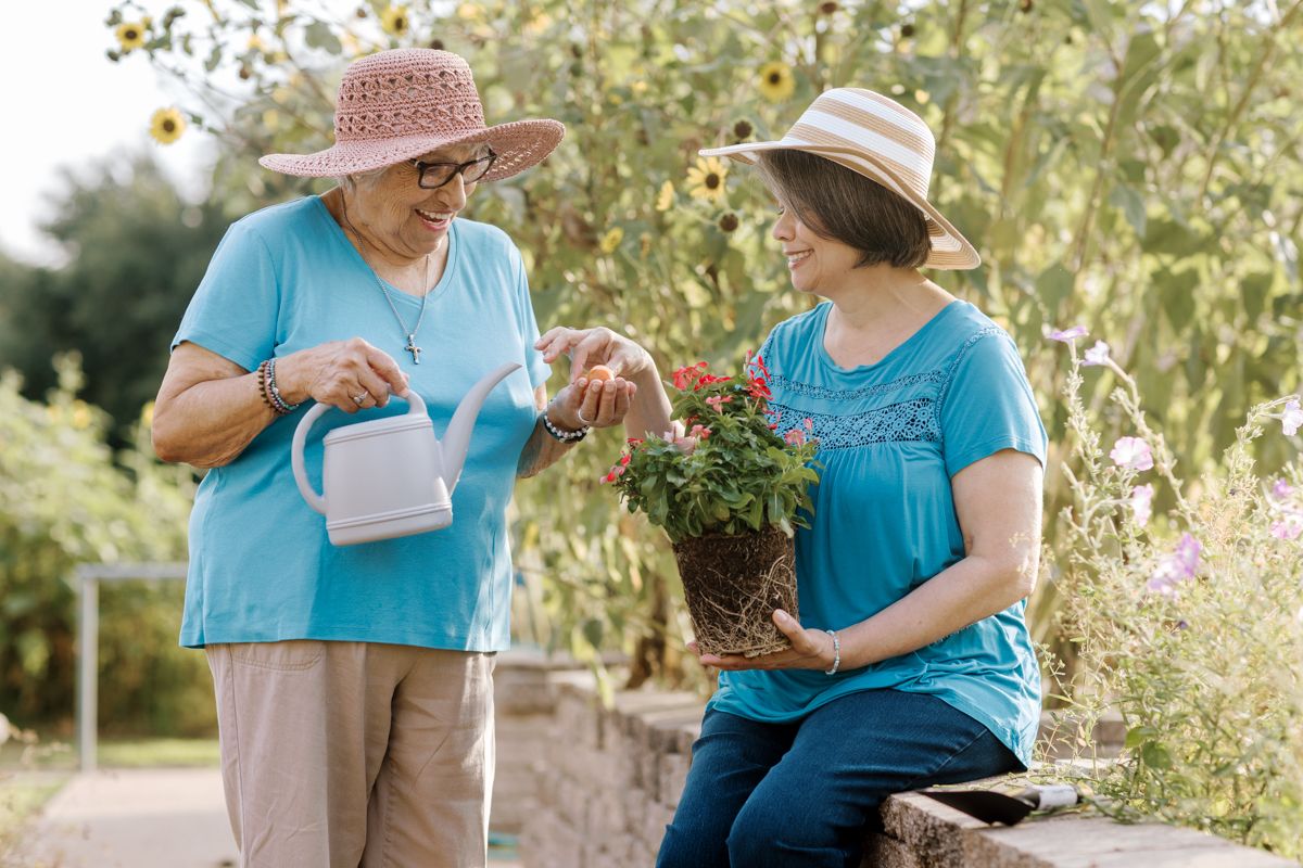Two women in sun hats garden together, one watering plants while the other holds a flowering pot outdoors.