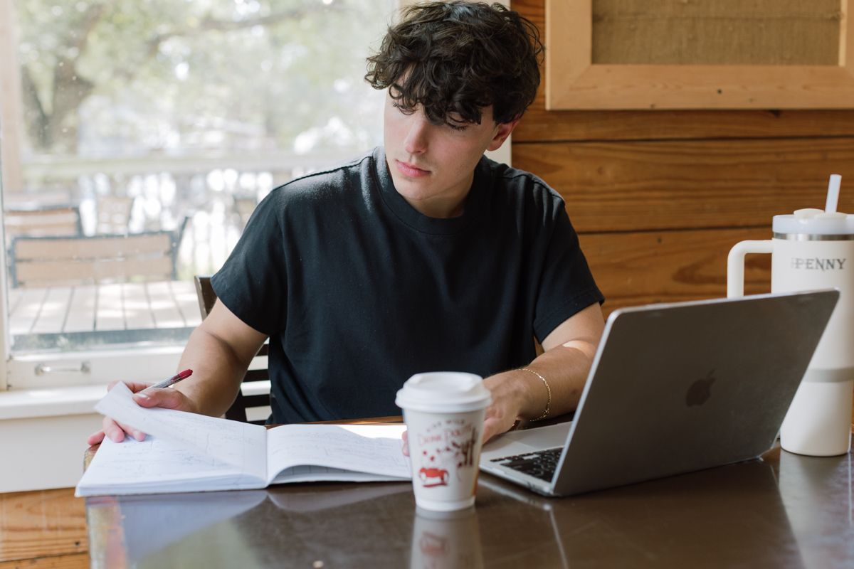 Young man studies with an open book and laptop at a cafe table with a to-go coffee cup.