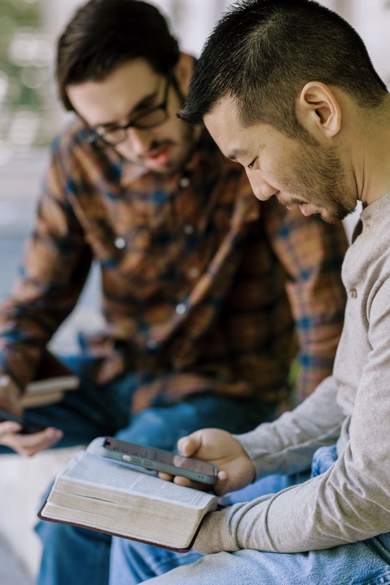 Two men studying the Bible together outdoors, one holding an open book and smartphone.