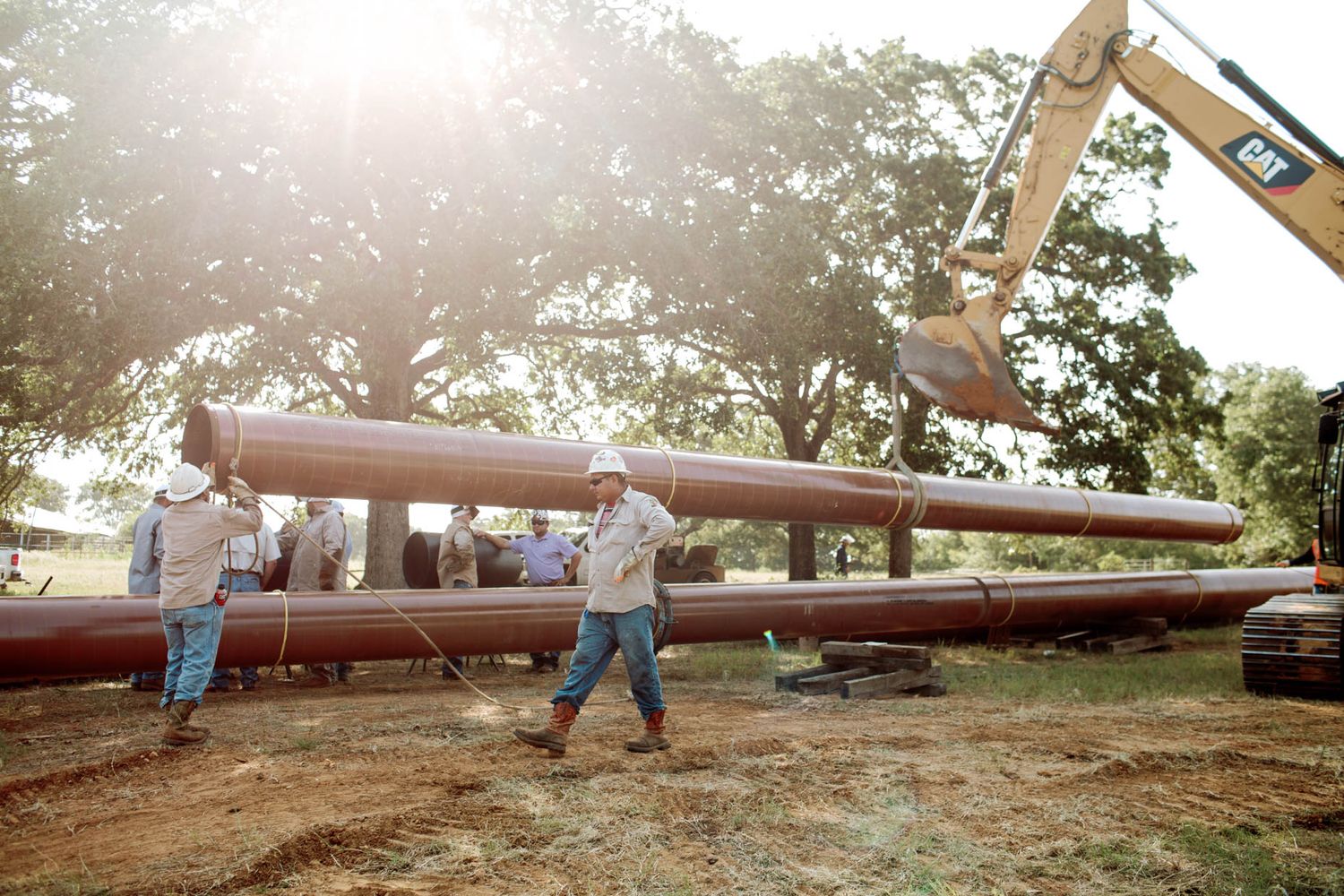 Workers and excavator handling large industrial pipes outdoors near trees on a construction site.