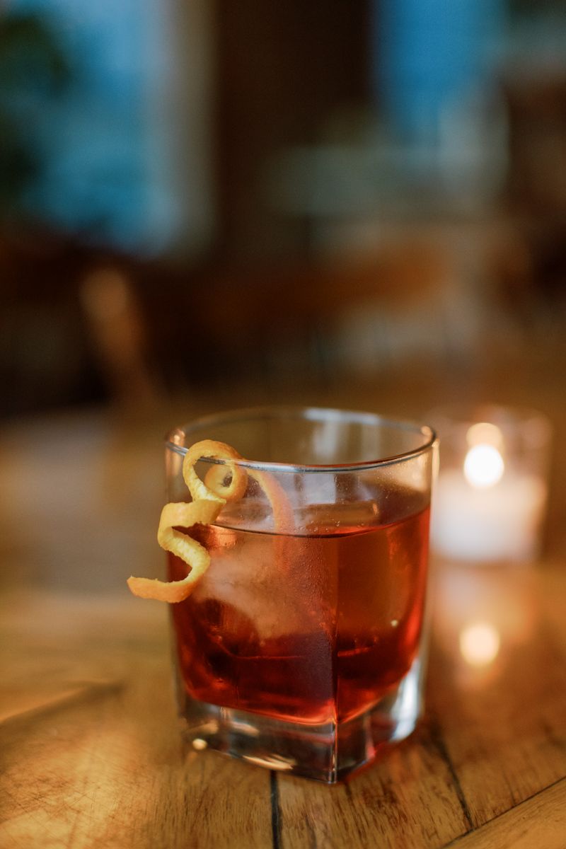 Classic cocktail with a citrus peel garnish served in a rocks glass beside a candle on a wooden bar.