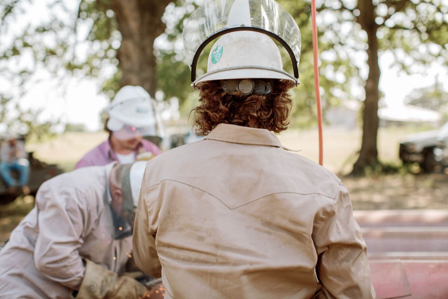 Two construction workers wearing white hard hats and beige work shirts at an outdoor job site.