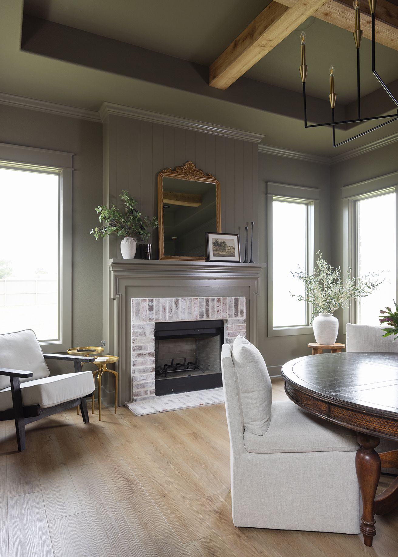 Moody olive green sitting room with brick fireplace, exposed wood beam, round dining table, and black chandelier.