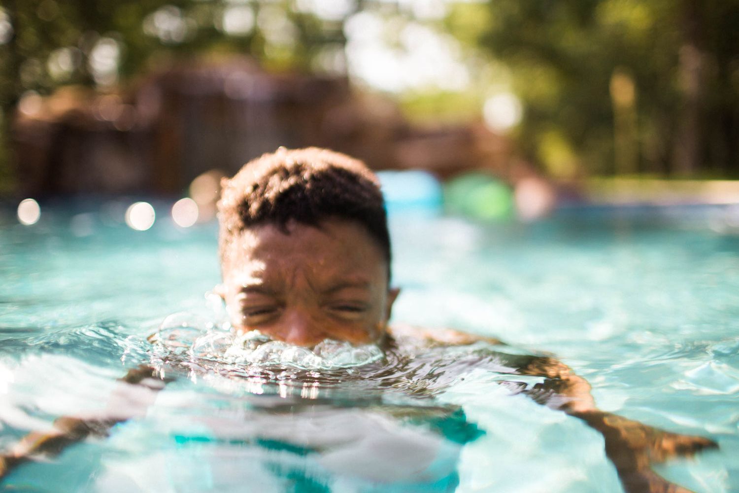 A young boy swimming in a clear blue outdoor pool on a sunny day, with his face partially submerged in water.