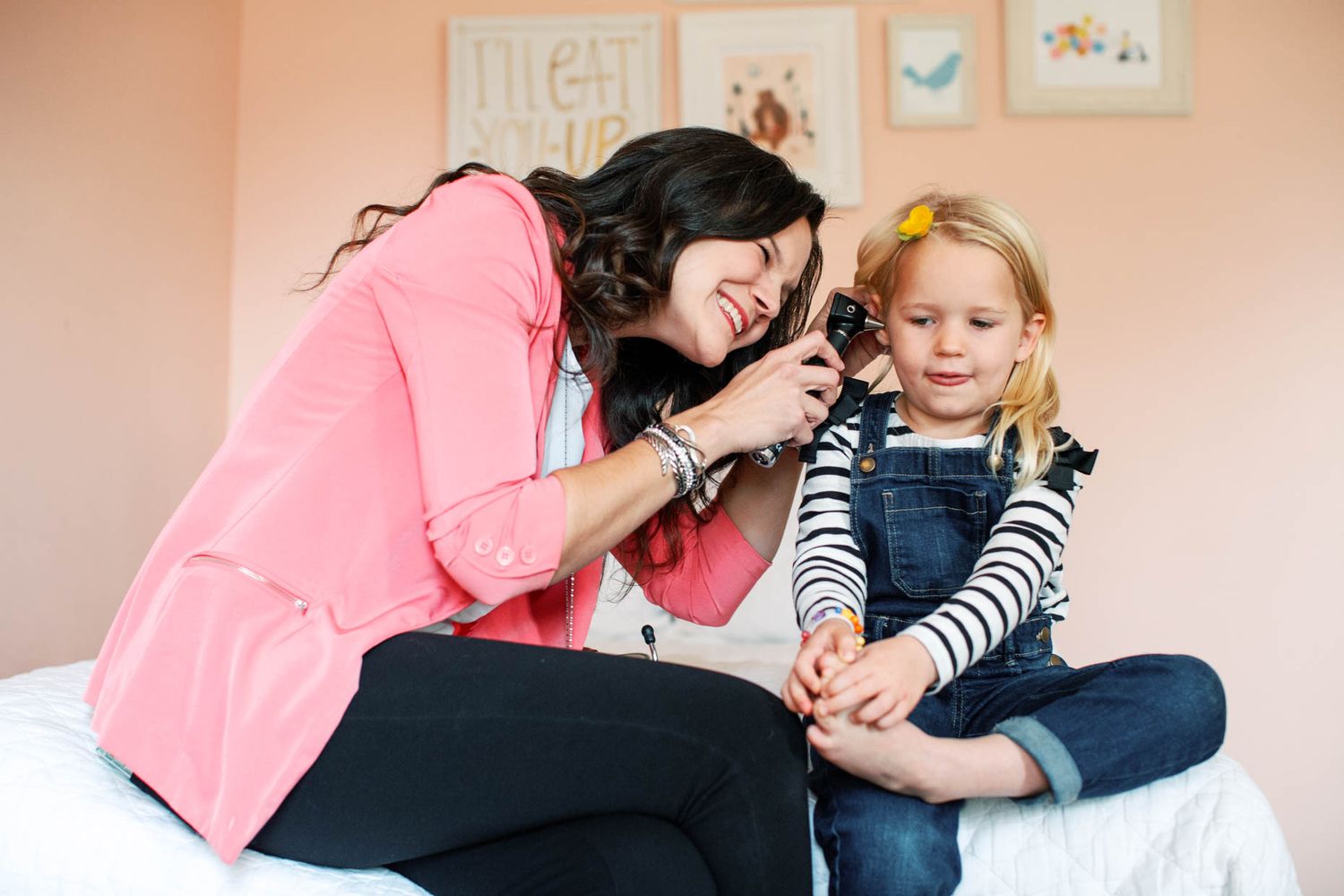 A smiling woman in pink examines a young blonde girl's ear with an otoscope while sitting on a bed.