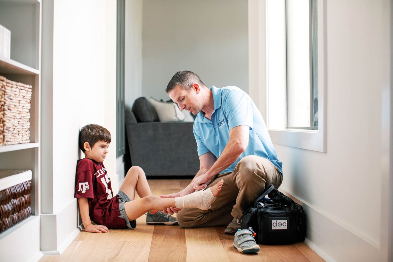 A medical professional in a blue polo examines a young boy's leg on a home's wooden floor.