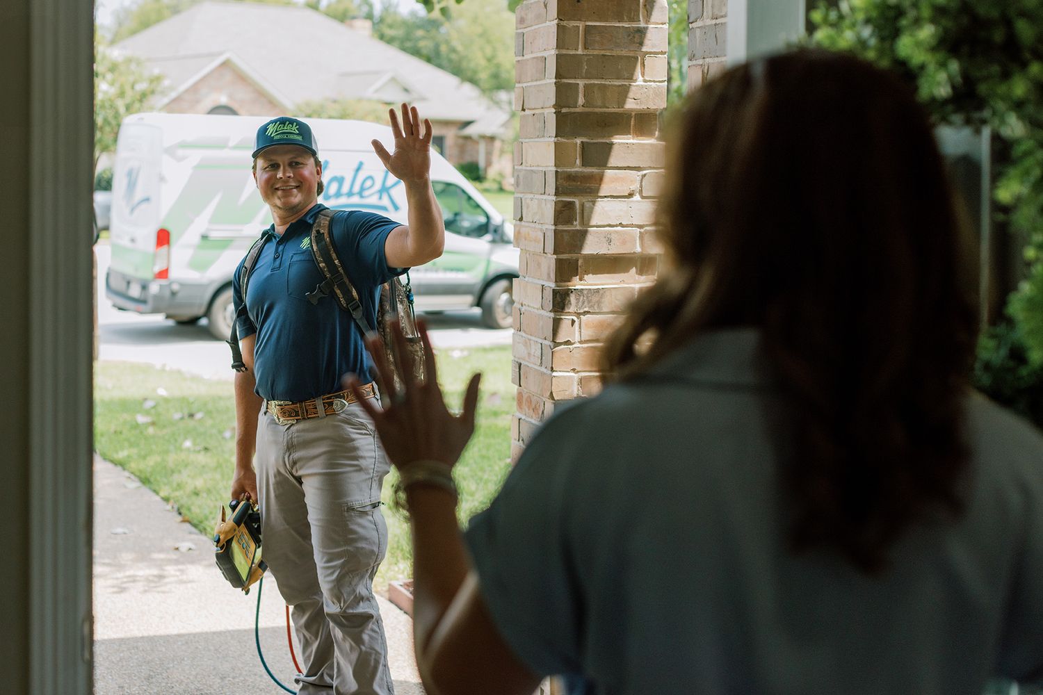 A smiling technician in a blue uniform waves goodbye to a homeowner at her front door after a service visit.