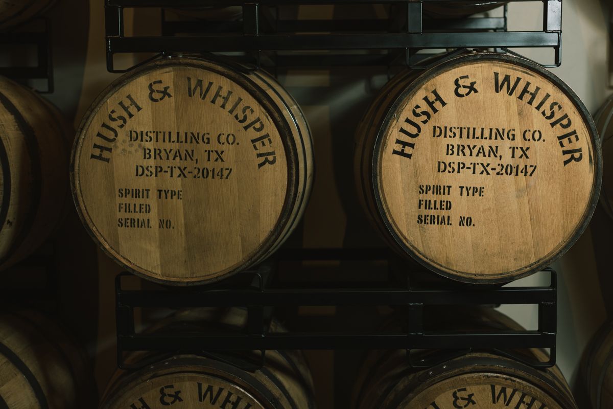 Wooden barrels labeled Hush and Whisper Distilling Co. Bryan TX stacked in a dimly lit warehouse.