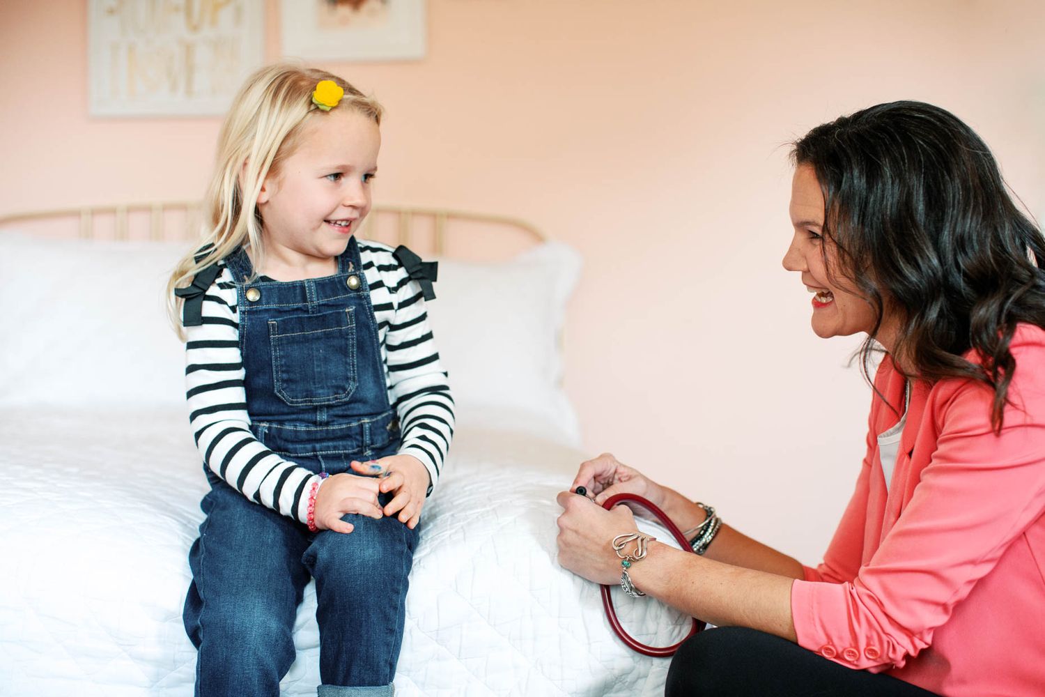 A pediatrician uses a stethoscope to examine a smiling young blonde girl in denim overalls during a home visit.