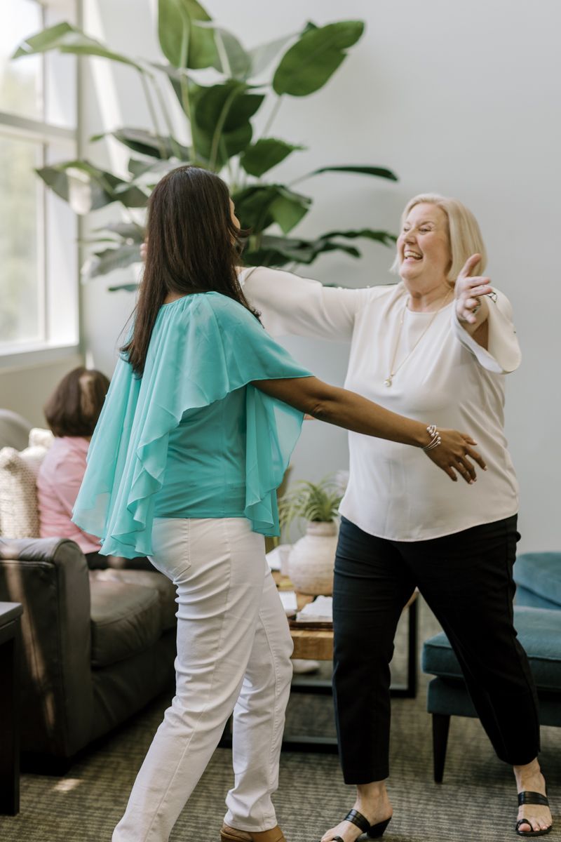 Two women dancing together joyfully in a bright living room with a large tropical plant in the background.